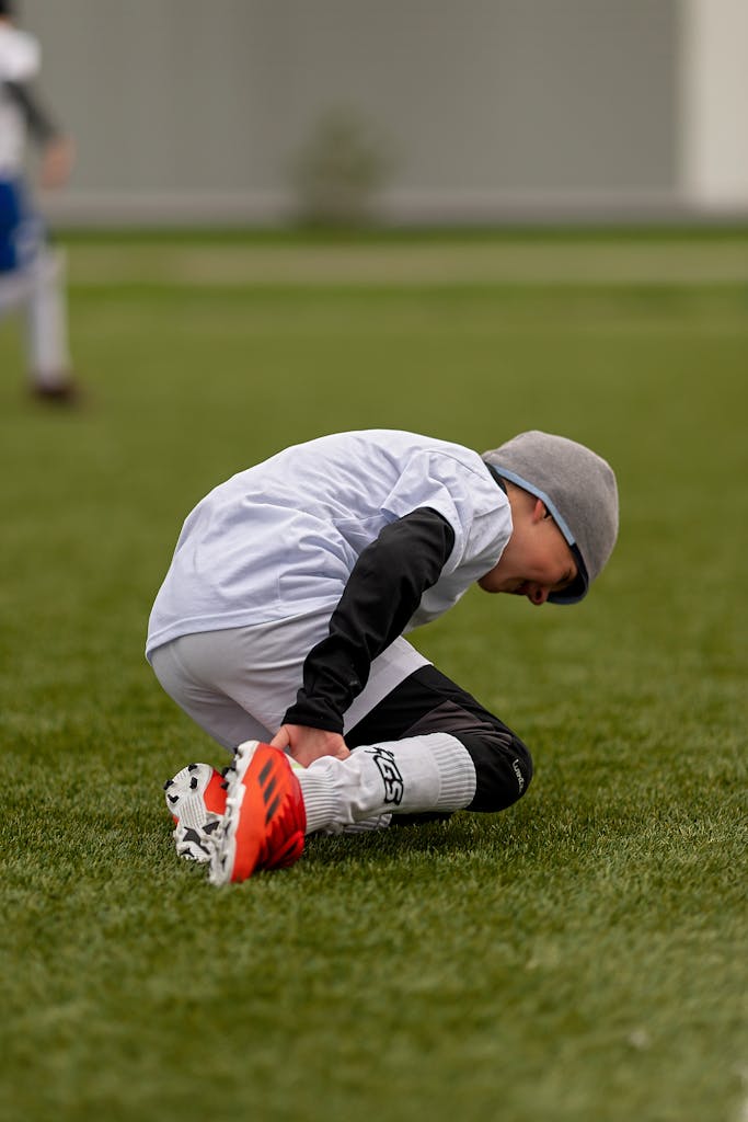 A young boy in sports gear kneeling on the grass during a soccer game outdoors.