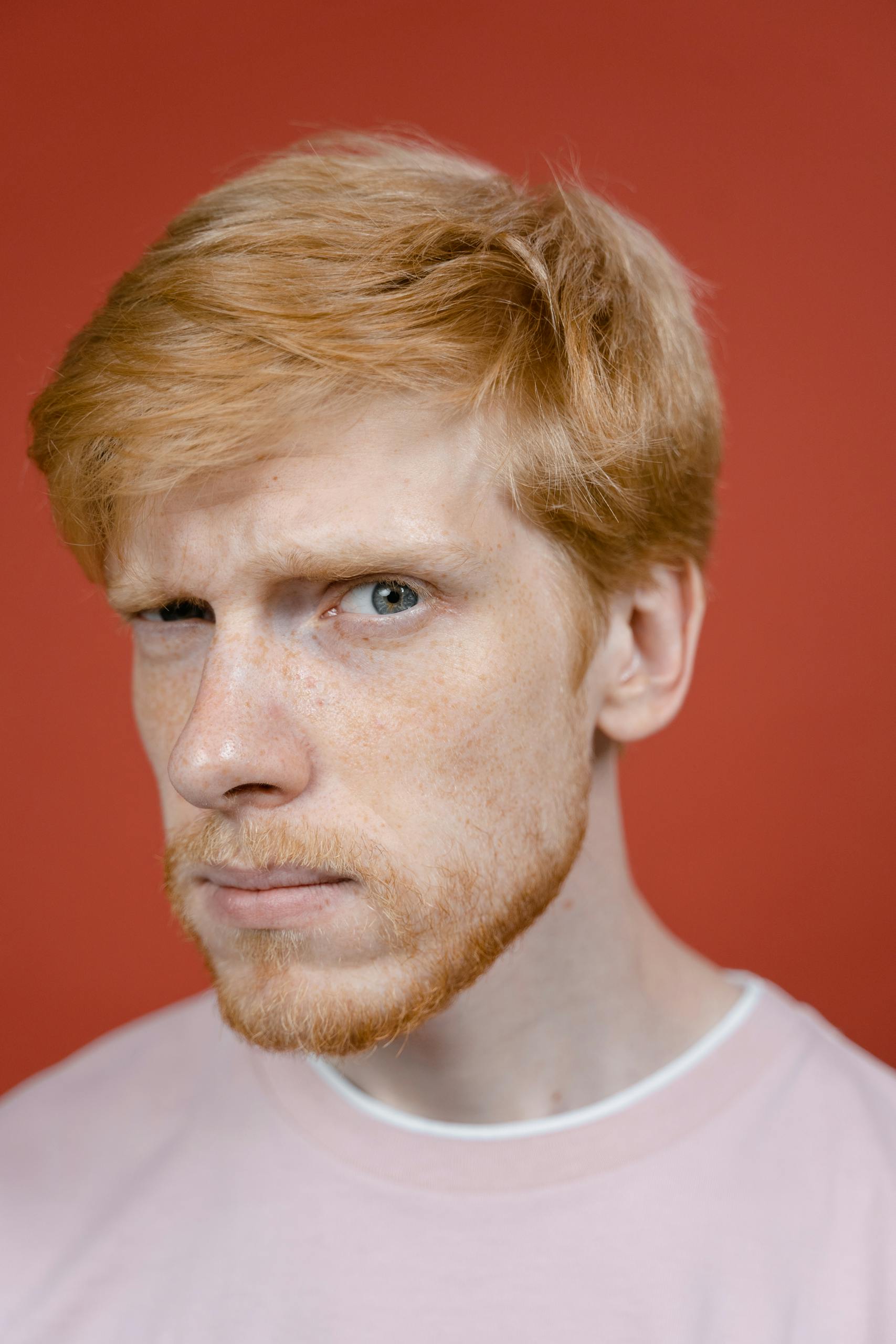 Close-up of a red-haired bearded man with a skeptical expression against a red backdrop.